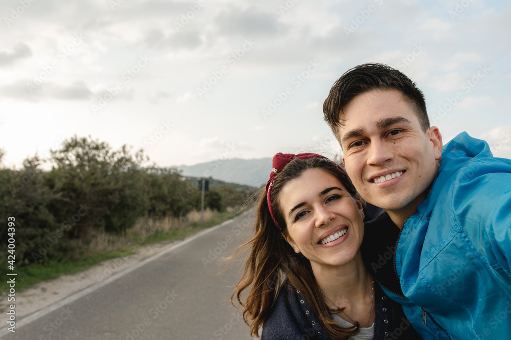 Young happy couple doing a selfie portrait smiling in the middle of a road. Traveling, trip, nomad concept