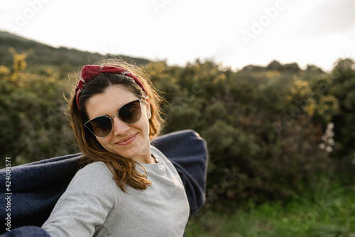 Happy casual young woman with sunglasses and scarf in countryside smiling, looking at the camera enjoying the landscape and fresh air.