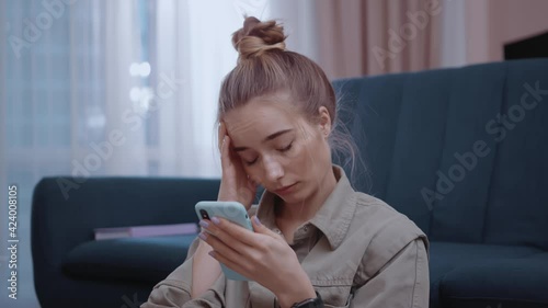 Portrait young woman use phone with headache sitting in home near sofa feel sick unhappy. Attractive, online, mobile, tired, cellphone, application, smartphone, internet. Slow motion