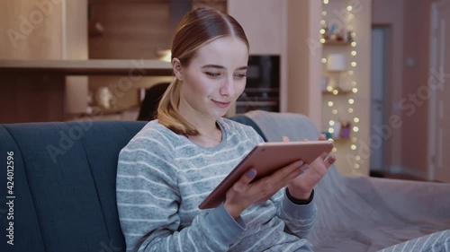 Smiling young woman sitting on couch using tablet computer at home evening. Relaxation, touchscreen, modern, reading, apartment, touching, convenience, paying, online, digital tablet, touchpad. Slow