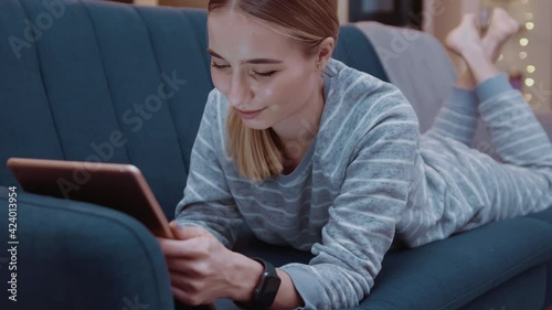 Cute young woman lying on couch using tablet computer at home evening. Relaxation, touchscreen, modern, reading, apartment, touching, convenience, paying, online, digital tablet, touchpad. Slow motion