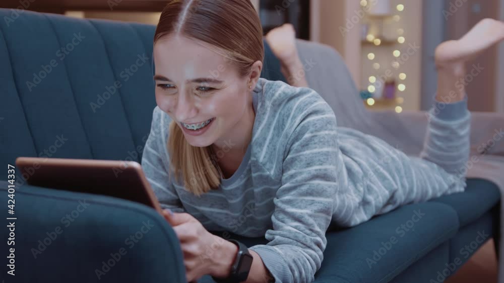 Happy young woman lying on couch using tablet computer at home evening. Relaxation, touchscreen, modern, reading, apartment, touching, convenience, paying, online, digital tablet, touchpad. Slow Happy young woman lying on couch using tablet computer at home evening. Relaxation, touchscreen, modern, reading, apartment, touching, convenience, paying, online, digital tablet, touchpad. Slow