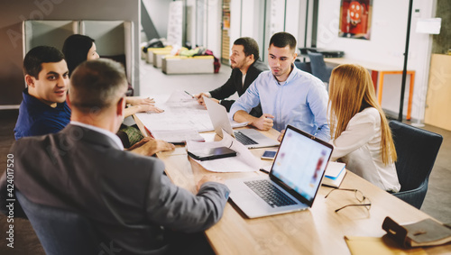 Group of productive employees collaborating at desktop in office discussing ideas for startup, male and female partners talking about trade and project analyzing details during briefing meeting © BullRun