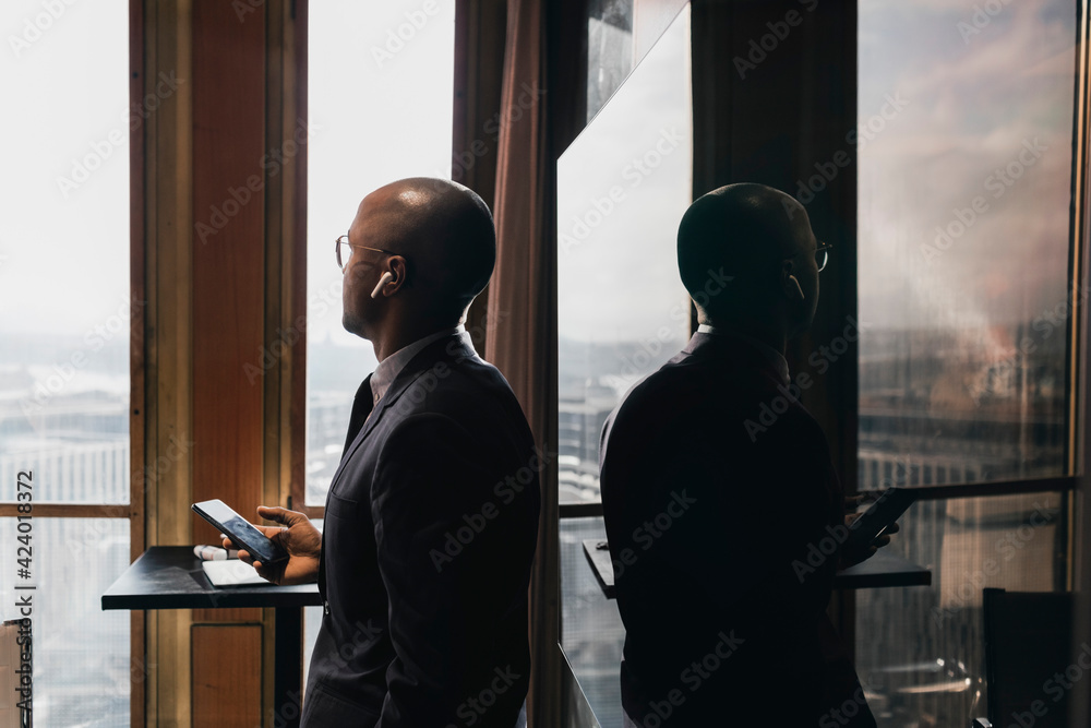 Side view of bald businessman wearing in-ear headphones holding smart phone looking away against television in board roo Side view of bald businessman wearing in-ear headphones holding smart phone looking away against television in board roo