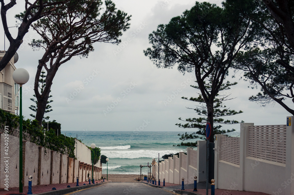 An asphalt road with two red sidewalks between two fences leads to the beach and turquoise sea with big waves in inclement weather
