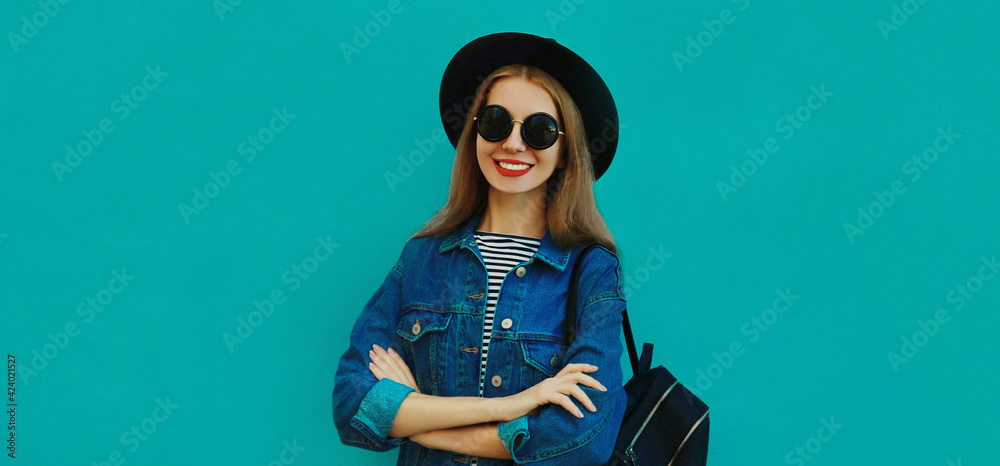 Portrait close up of smiling young woman wearing a black round hat, denim jacket and backpack on a blue background Portrait close up of smiling young woman wearing a black round hat, denim jacket and backpack on a blue background