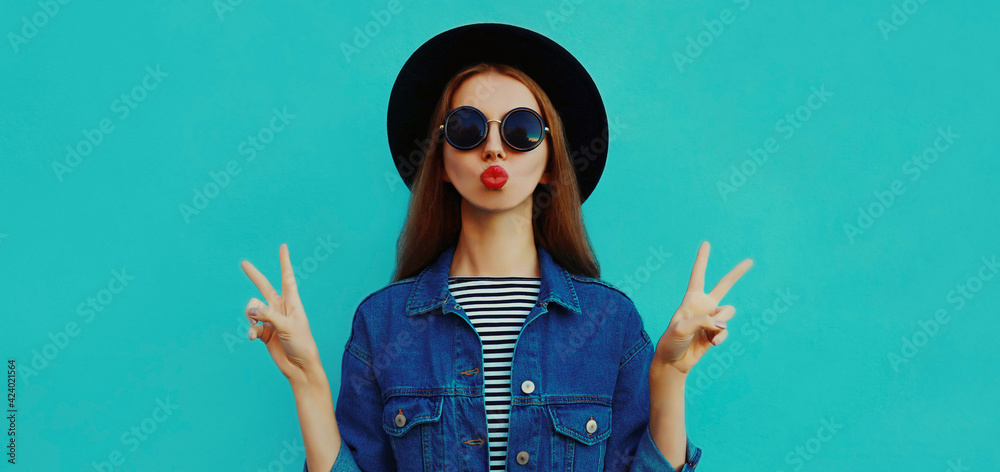 Portrait close up of young woman blowing her red lips sending sweet air kiss wearing a black round hat, denim jacket on a blue background Portrait close up of young woman blowing her red lips sending sweet air kiss wearing a black round hat, denim jacket on a blue background