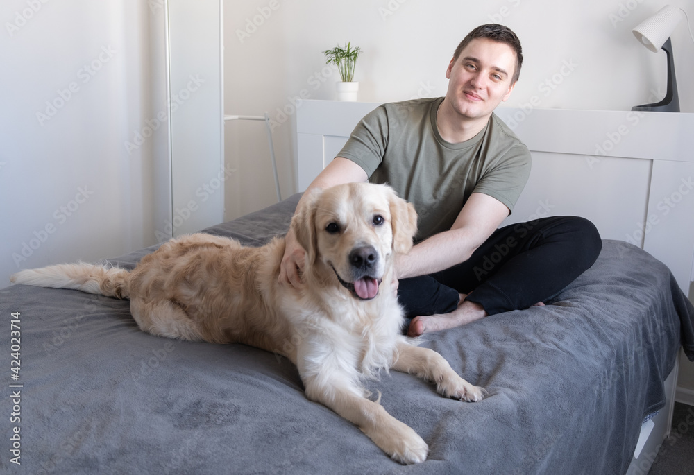 A young man is playing on the bed with a dog. Guy with companion golden retriever sitting at home