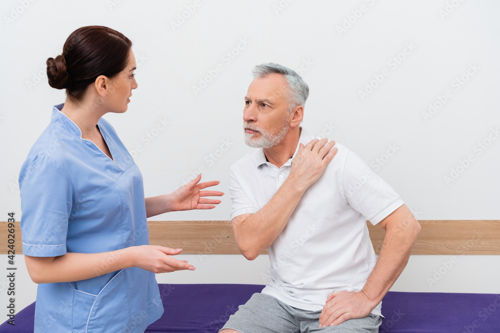 Fototapeta premium rehabilitologist gesturing while talking to mature man touching shoulder in clinic