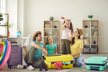 Happy family watching one of excited little daughter playing with toy plane sitting on floor with packed belongings in travel suitcase around. Rejoice of travel opportunity after quarantine lockdown