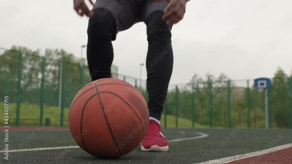 Close up shot with focus of ball lying on ground on outdoor basketball court. Black man in hoodie running towards camera and picking ball up, then dribbling it Close up shot with focus of ball lying on ground on outdoor basketball court. Black man in hoodie running towards camera and picking ball up, then dribbling it