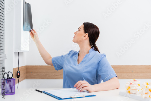 brunette rehabilitologist looking at x-ray while sitting near clipboard photo