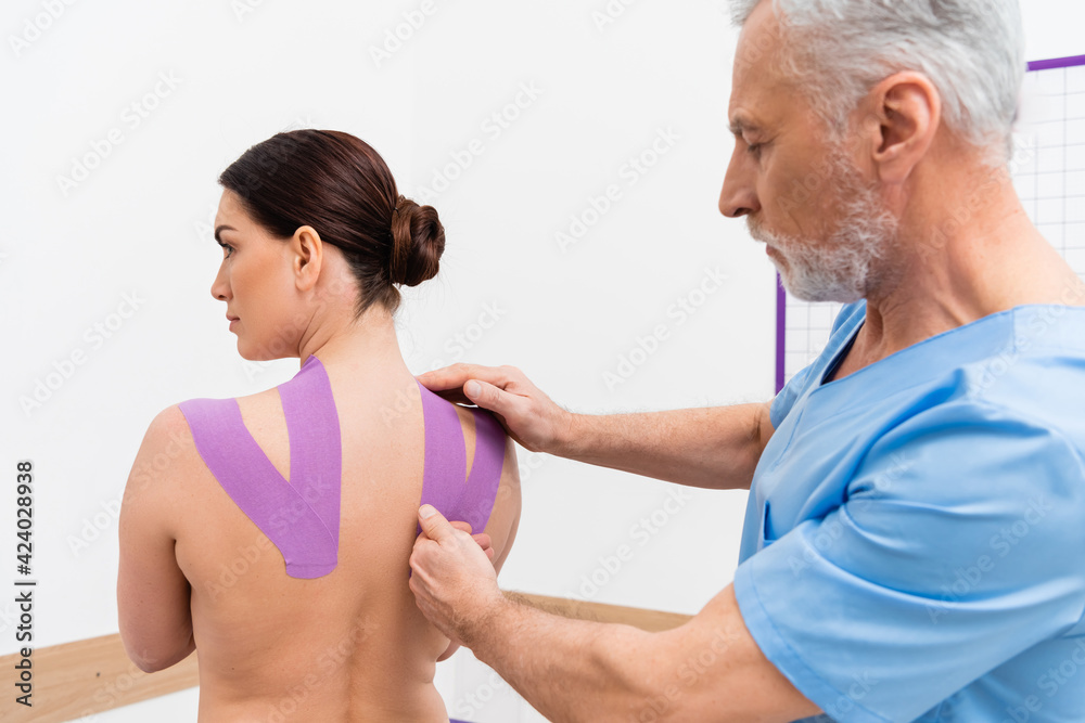 mature rehabilitologist applying tape on back of woman during kinesiotherapy mature rehabilitologist applying tape on back of woman during kinesiotherapy