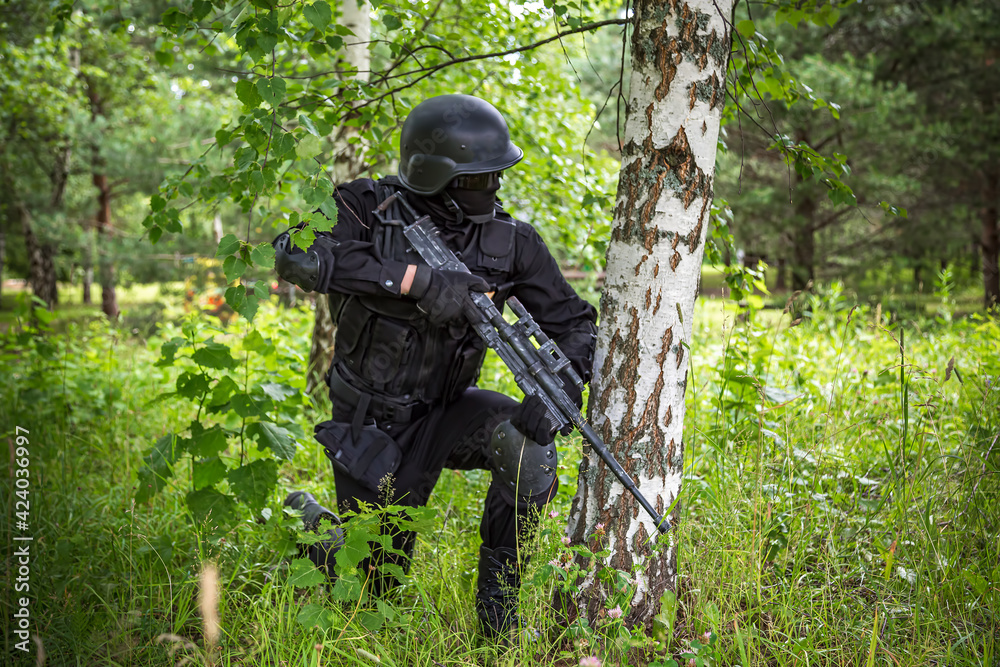 A soldier in special black equipment. Warrior without insignia and stripes. Special forces in the woods aiming with weapons. Tactical exercises.