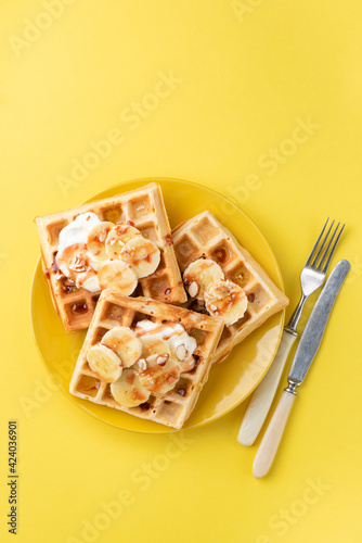 Waffles with banana, almond slices, cream and caramel sauce isolated on yellow background, top view. Sweet breakfast or dessert waffles photo