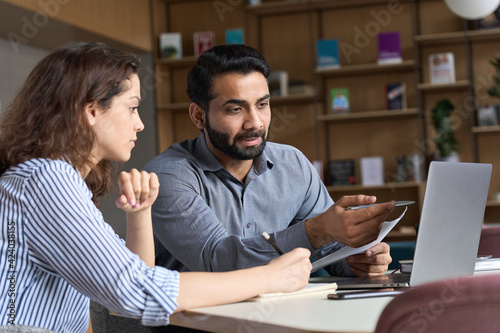 Professional indian teacher, executive or mentor helping latin student, new employee, teaching intern, explaining online job using laptop computer, talking, having teamwork discussion in office.