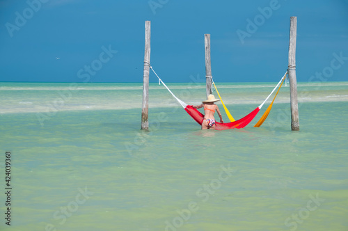 A young woman in a sun hat and bikini relaxes in a hammock by the sea on Holbox Island in Mexico. In the background the cloudless blue sky and the Caribbean ocean. Concept travel vacations