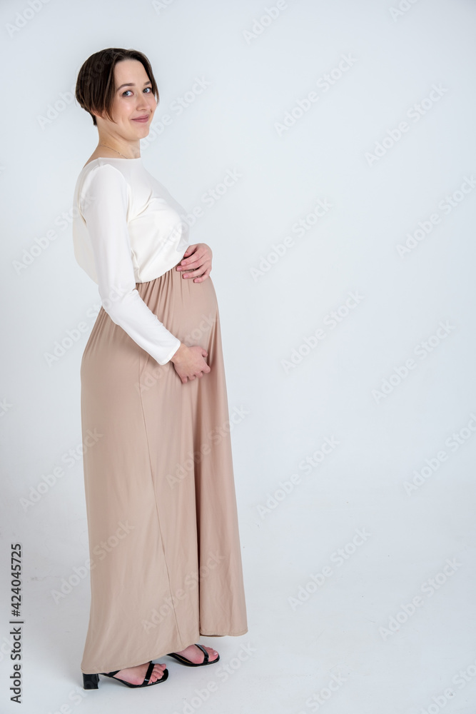 Studio portrait of elegant young adult pregnant woman in white beige dress on white background, happy pregnancy concept Studio portrait of elegant young adult pregnant woman in white beige dress on white background, happy pregnancy concept