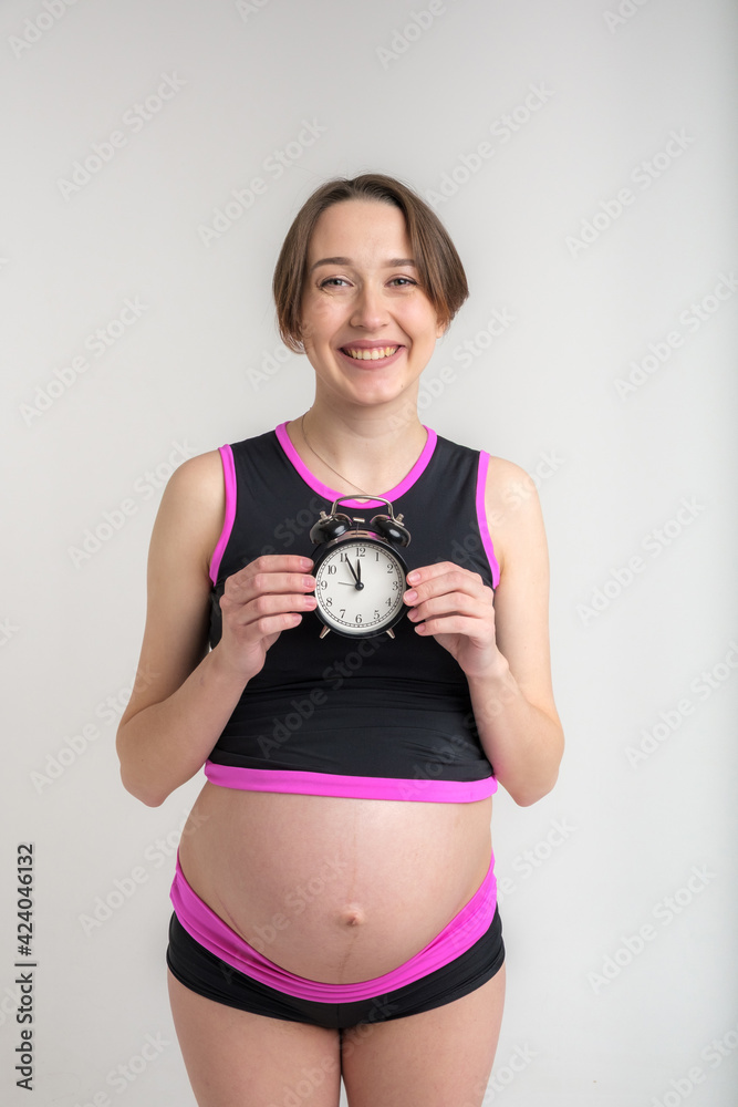Young adult pregnant girl in sportswear with alarm clock. Time for sports concept. Yoga and fitness in anticipation of childbirth. White background. Young adult pregnant girl in sportswear with alarm clock. Time for sports concept. Yoga and fitness in anticipation of childbirth. White background.