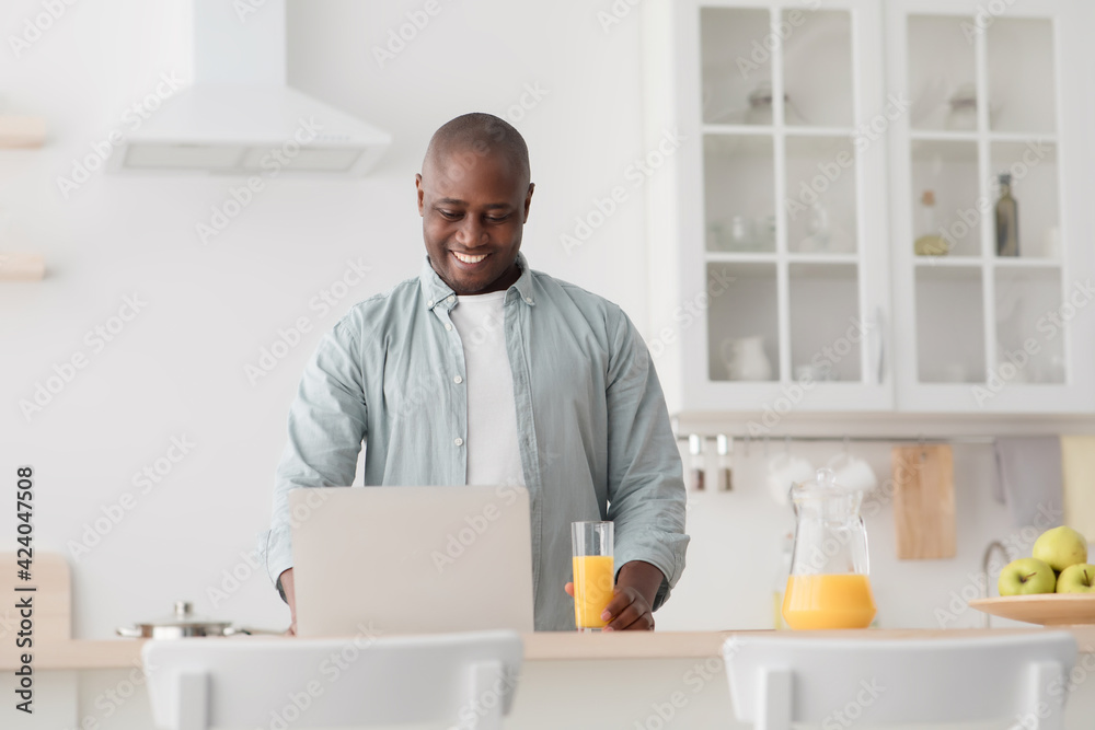 Happy african american mature man drinking orange juice and watching movie on laptop in kitchen