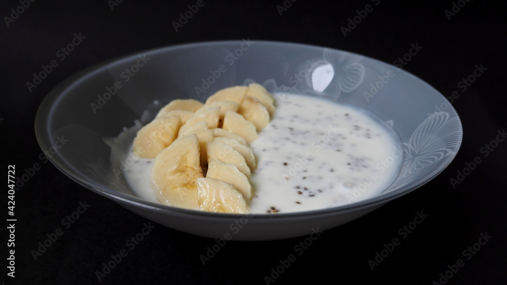 yogurt with sliced banana slices and chia seeds stands in a gray round plate on a black background. side view . natural snack food yogurt with sliced banana slices and chia seeds stands in a gray round plate on a black background. side view . natural snack food