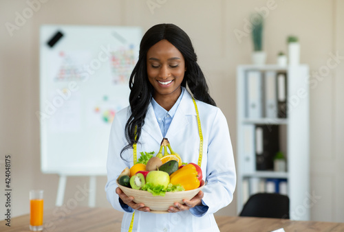 Portrait of joyful black nutritionist in lab coat holding bowl of fresh fruits and veggies at weight loss clinic
