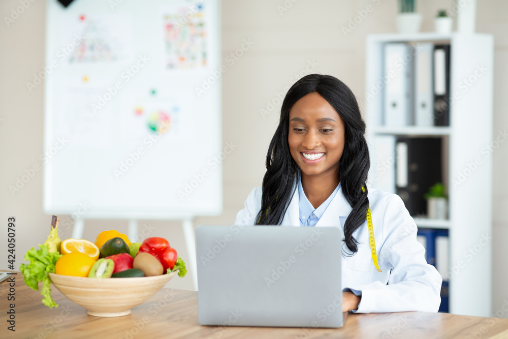 Cheerful black female dietitian using laptop pc, creating online weight loss program at modern clinic, free space Cheerful black female dietitian using laptop pc, creating online weight loss program at modern clinic, free space