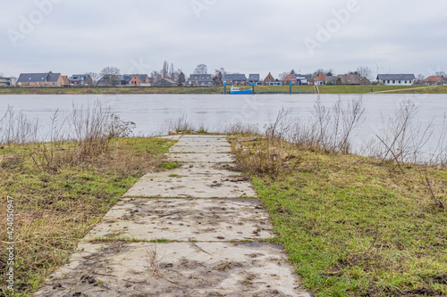 Small asphalt path heading to the river Maas among sparse green grass on the Dutch side, a Belgian village on the other side of the river, cloudy day in Geulle in South Limburg, Netherlands photo