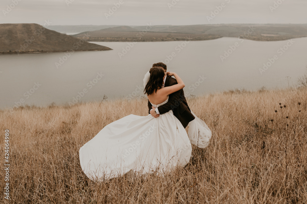 A loving couple wedding newlyweds in a white dress and a suit walk hug kissing on the tall grass in the summer autumn field on the mountain above the river. sunset. bakota ukraine A loving couple wedding newlyweds in a white dress and a suit walk hug kissing on the tall grass in the summer autumn field on the mountain above the river. sunset. bakota ukraine