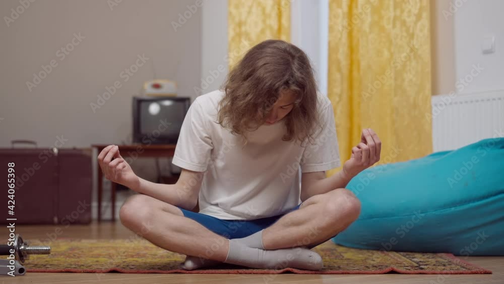 Portrait of calm young retro man with long curly hair falling asleep sitting in lotus pose indoors. 1990s guy practicing yoga exercise at home. Yogi meditating in 1980s. Portrait of calm young retro man with long curly hair falling asleep sitting in lotus pose indoors. 1990s guy practicing yoga exercise at home. Yogi meditating in 1980s.
