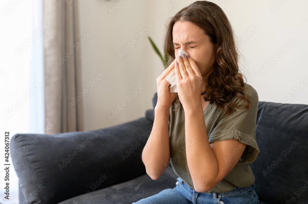 Sick young woman sneezing into handkerchief or tissue at home, feeling unwell from flu or allergy, female has running nose and other symptoms of viral diseases, covid-19 Sick young woman sneezing into handkerchief or tissue at home, feeling unwell from flu or allergy, female has running nose and other symptoms of viral diseases, covid-19