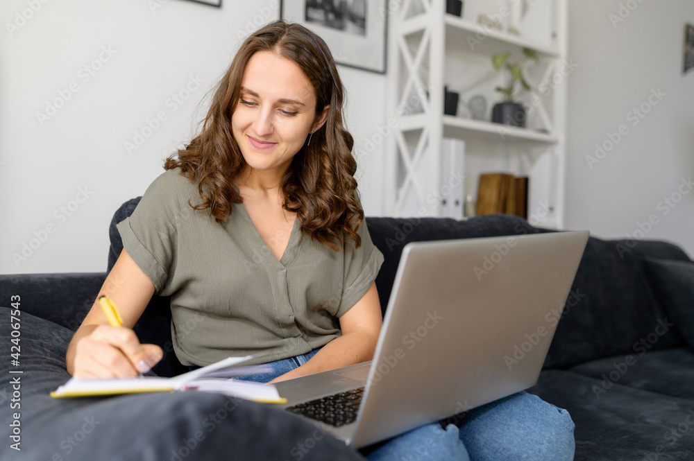 Focused female student studying on the distance, using laptop for watching online lectures, woman taking notes with a pen sitting on the couch at home, female employee working remotely Focused female student studying on the distance, using laptop for watching online lectures, woman taking notes with a pen sitting on the couch at home, female employee working remotely