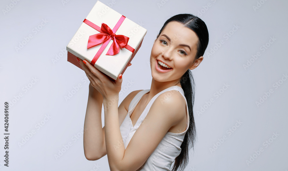 Cheerful woman holding present box in studio on gray background. Smiling beautiful young girl expressing happy emotions. Seasonal discounts and sale. Cheerful woman holding present box in studio on gray background. Smiling beautiful young girl expressing happy emotions. Seasonal discounts and sale.