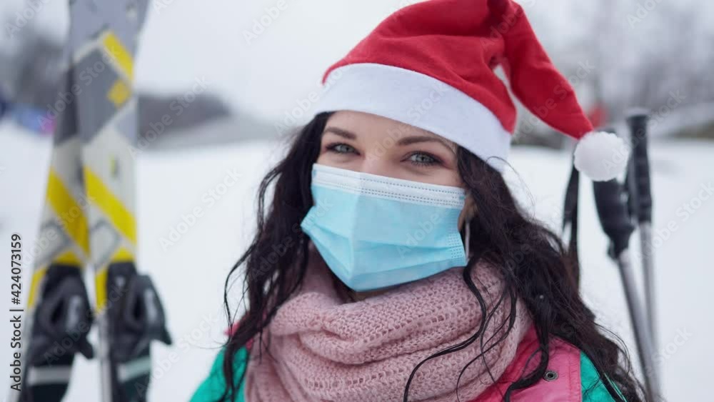 Headshot of joyful smiling young woman in Covid face mask and Christmas hat looking at camera. Close-up of happy relaxed tourist enjoying winter leisure outdoors in snowy mountains Headshot of joyful smiling young woman in Covid face mask and Christmas hat looking at camera. Close-up of happy relaxed tourist enjoying winter leisure outdoors in snowy mountains