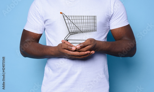 Black man with white t-shirt holds a shopping cart. cyan background © alphaspirit