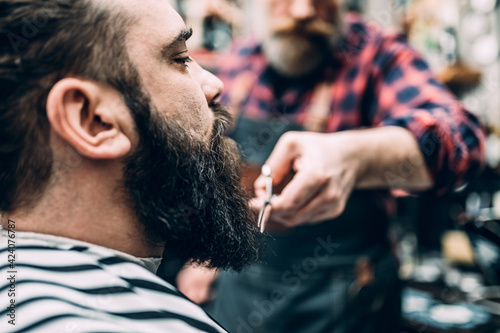 The barber trims the beard of the customer in his barber shop. Trendy and stylish beard styling and cut.