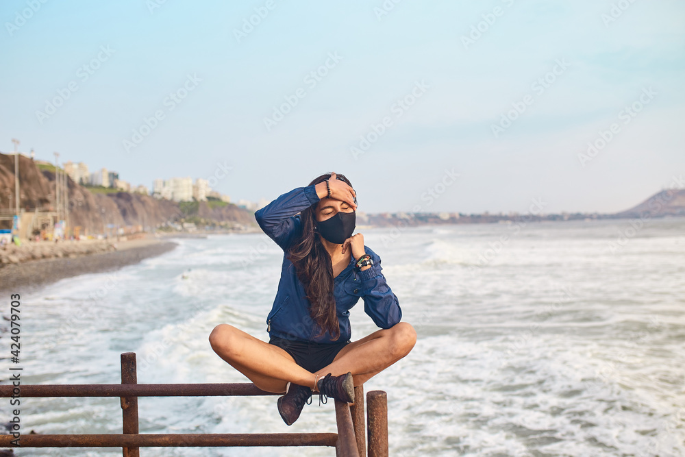 mujer joven con mascarilla feliz en la playa. Momento de relajacion a orillas del mar durante un atardecer. Fondo de olas y mar
