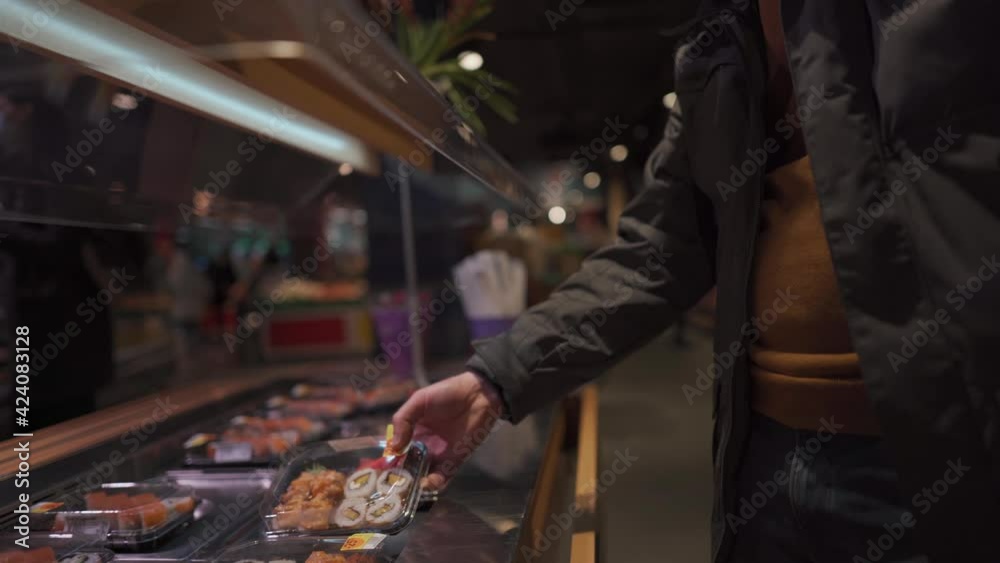 Man shopper in mask chooses sushi in plastic box on shelf in supermarket. Japanese takeaway food. No mask, no entrance. Prepared healthy food in disposable plastic container. Lunch box with sushi Man shopper in mask chooses sushi in plastic box on shelf in supermarket. Japanese takeaway food. No mask, no entrance. Prepared healthy food in disposable plastic container. Lunch box with sushi