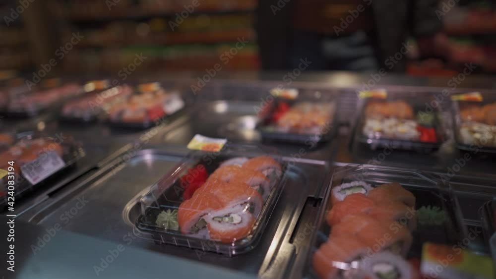 Man shopper in mask chooses sushi in plastic box on shelf in supermarket. Japanese takeaway food. No mask, no entrance. Prepared healthy food in disposable plastic container. Lunch box with sushi Man shopper in mask chooses sushi in plastic box on shelf in supermarket. Japanese takeaway food. No mask, no entrance. Prepared healthy food in disposable plastic container. Lunch box with sushi