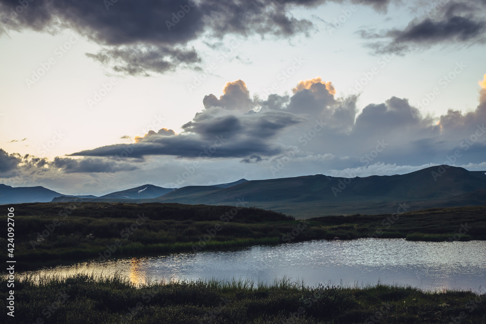 Fototapeta premium Scenic sunset landscape with lake under vivid orange gradient sky. Colorful mountain sunrise scenery with illuminating color in cloudy sky and golden reflection on lake. Clouds in gradient dawn sky.