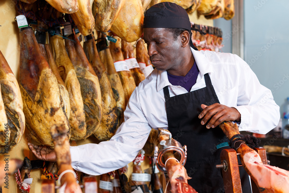 Confident butcher working behind counter in delicatessen shop, checking quality of jamon