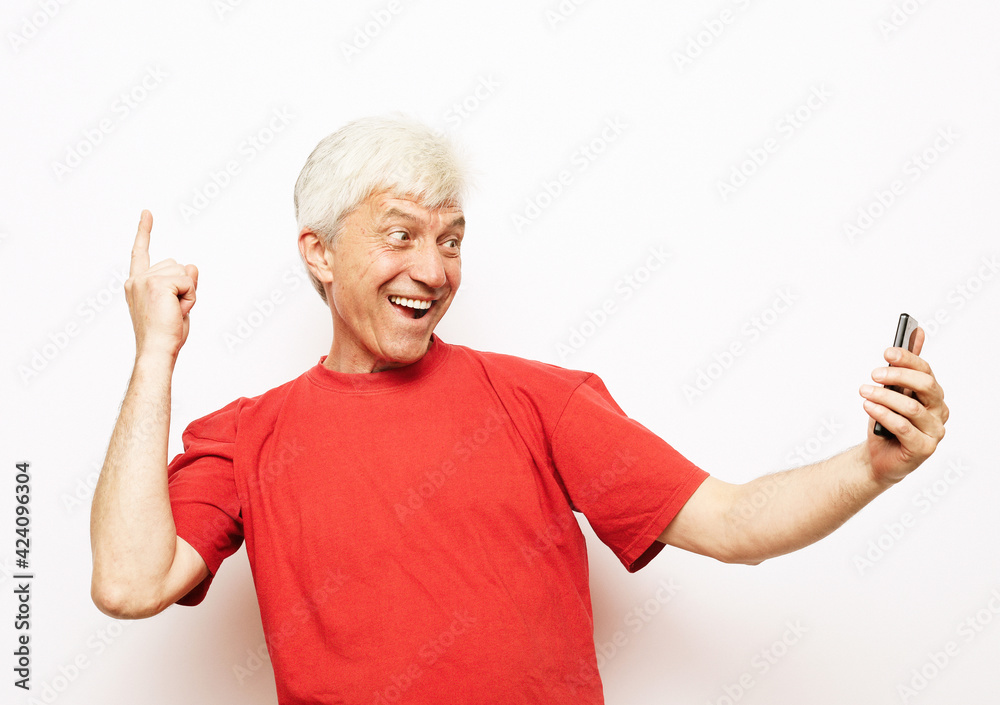 Senior man wearing red t-shirt smiling and happy, taking a selfie, holding the camera, cheerful expression Senior man wearing red t-shirt smiling and happy, taking a selfie, holding the camera, cheerful expression