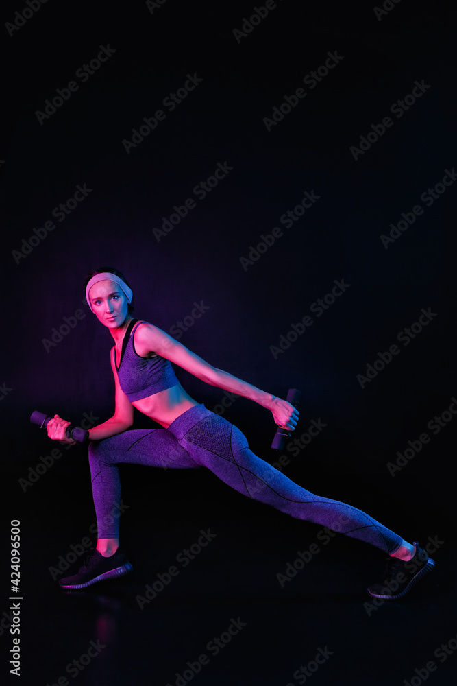 A slim, fit girl is stretching on a black background in a mixed color light. Self-improvement and fitness exercises for women. Copy space neon trend