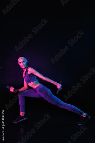A slim, fit girl is stretching on a black background in a mixed color light. Self-improvement and fitness exercises for women. Copy space neon trend