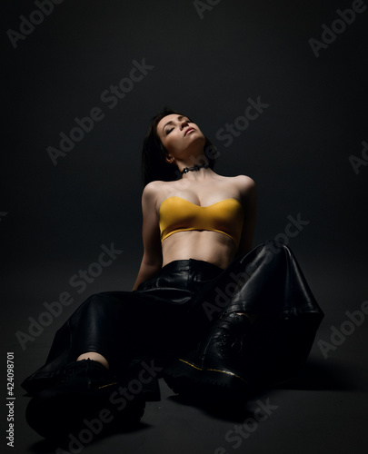 Young sensual languid brunette woman in leather pants, brutal choes and bra sits on floor with head thrown back and eyes closed over black background. Fashion, stylish casual look for woman concept