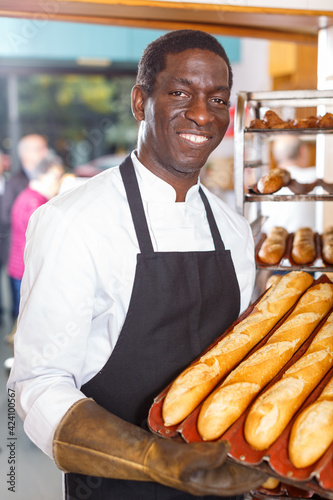 Portrait of successful owner of bakery holding tray with fresh baguettes..