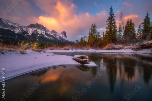 Three Sisters from Policeman Creek after sunrise reflected in the Bow River. The Three Sisters are a trio of peaks near Canmore, Alberta, Canada. They are known as Big, Middle and little Sister © Han