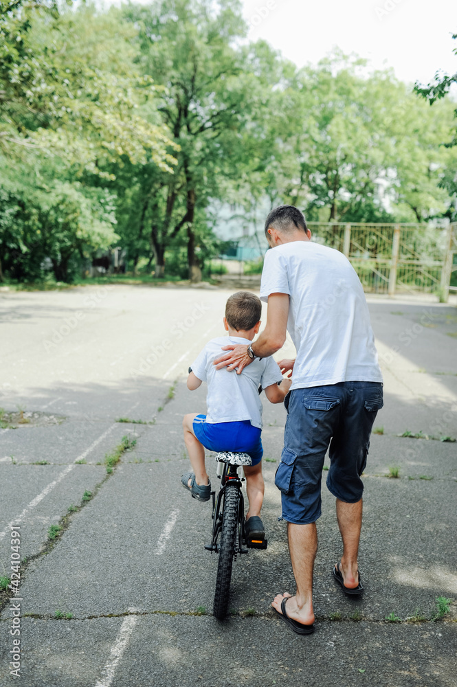 father teaches preschooler son to ride two-wheeled bicycle by holding him behind back. parental assistance to children. learning to ride adult bicycle. vertical, selective focus