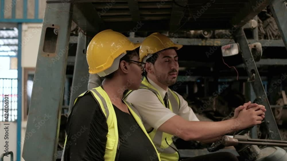 Caucasian male and black female engineers in safety uniforms and helmets on the forklift. Two employees taking about stock order of auto spare parts, engines, and machines in the factory warehouse. Caucasian male and black female engineers in safety uniforms and helmets on the forklift. Two employees taking about stock order of auto spare parts, engines, and machines in the factory warehouse.