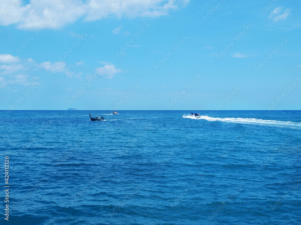 View from the water to the moving speedboat and longtail boats. Small rocky island on the distance. Blue sea, white clouds on the sky. Thailand tropical resort, boat trip, cruise. Seascape, ocean.